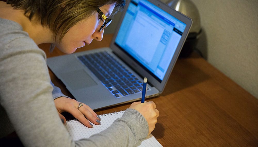 Student writing on notepad with laptop