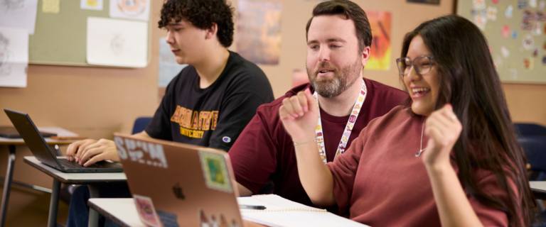 A classroom scene featuring a male teacher with a short beard and a lanyard around his neck, leaning in to assist a female student sitting in the foreground. The female student wears glasses and a maroon top, smiling joyfully with raised hands near her sticker-covered laptop. In the background, another male student in a black Arizona State University t-shirt sits at a desk typing on his own laptop.