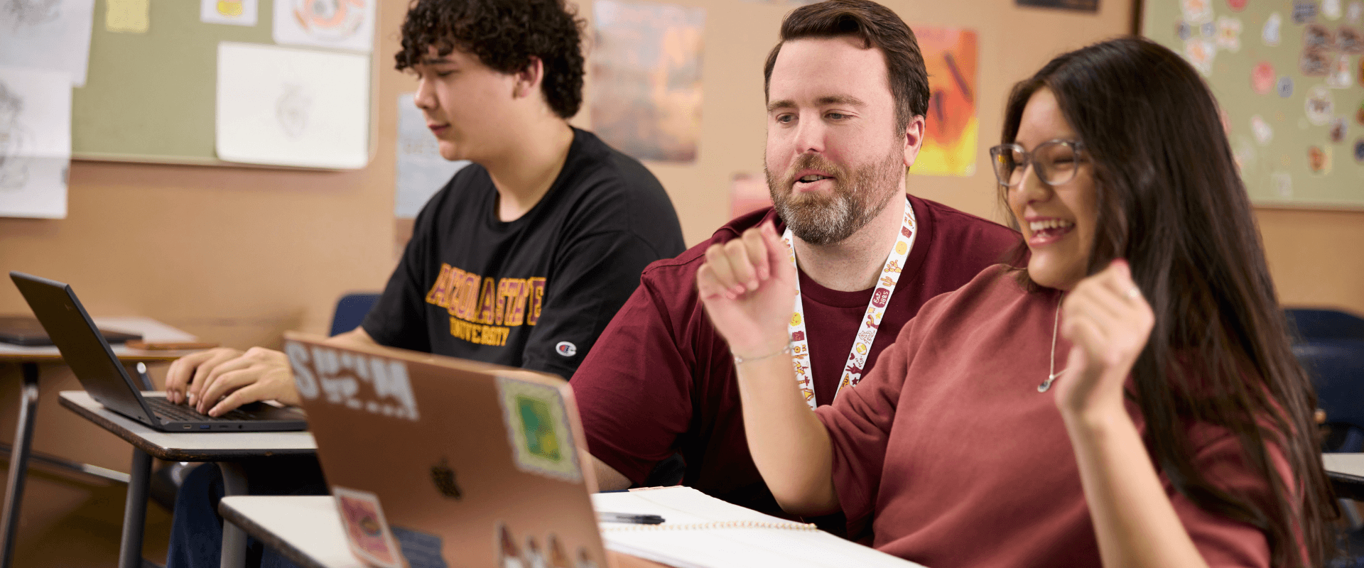 A classroom scene featuring a male teacher with a short beard and a lanyard around his neck, leaning in to assist a female student sitting in the foreground. The female student wears glasses and a maroon top, smiling joyfully with raised hands near her sticker-covered laptop. In the background, another male student in a black Arizona State University t-shirt sits at a desk typing on his own laptop.