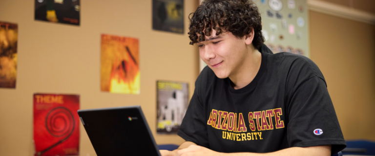 A young man with dark, curly hair wearing a black Arizona State University t-shirt looking down at a open laptop screen in a room with posters on the wall.