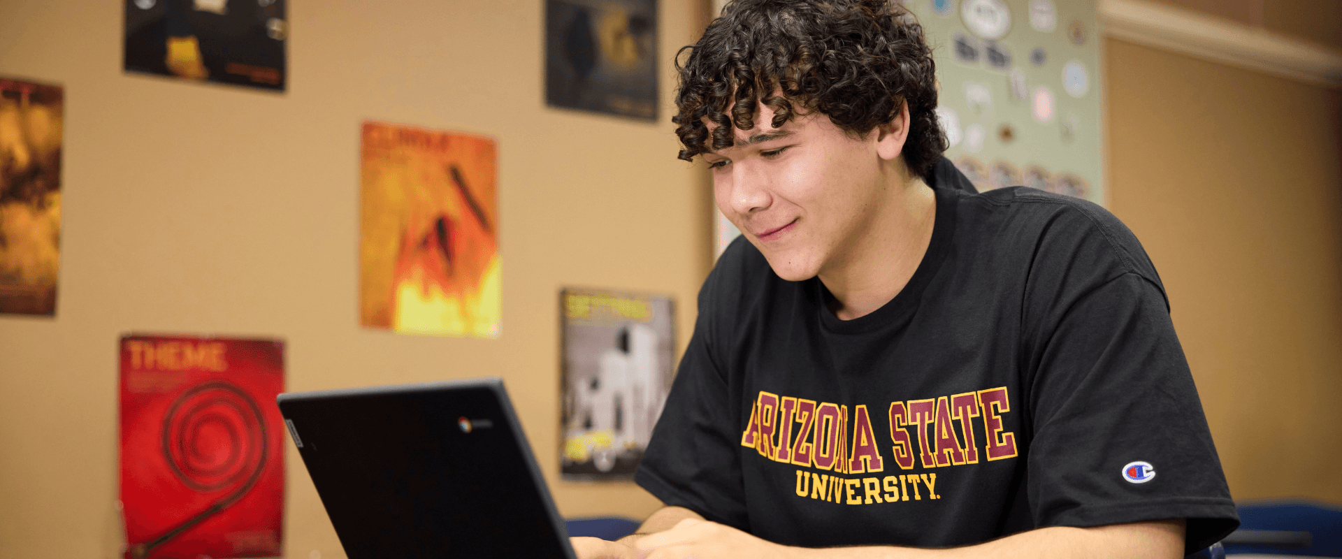 A young man with dark, curly hair wearing a black Arizona State University t-shirt looking down at a open laptop screen in a room with posters on the wall.